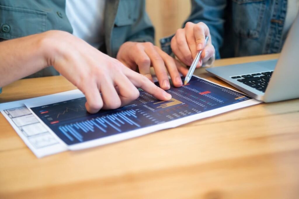 Two people reviewing financial charts and data on a printed report, pointing with fingers and a pen, with a laptop open on a wooden desk.