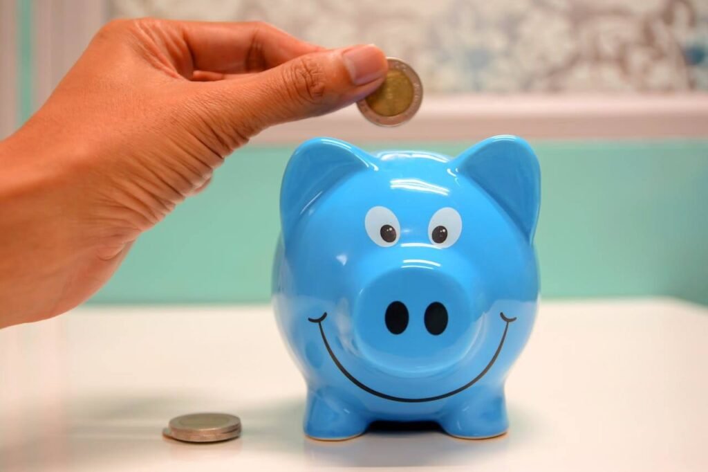 Hand dropping a coin into a smiling blue piggy bank, symbolizing saving money and building financial habits.