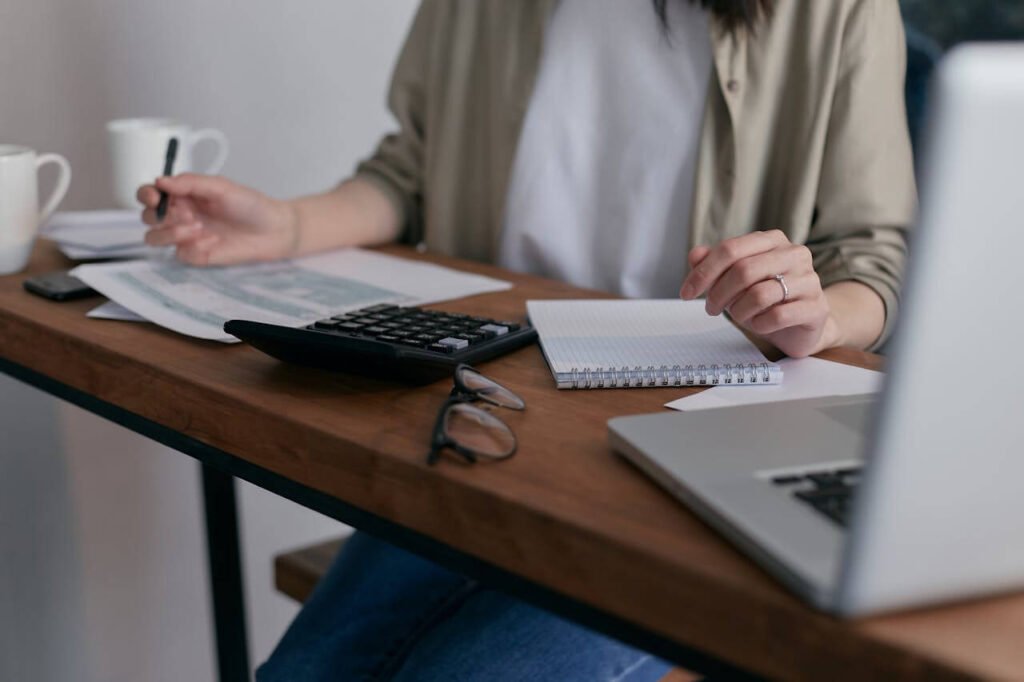 Person sitting at a desk working with financial documents, a calculator, notebook, glasses, and a laptop, with coffee mugs in the background.