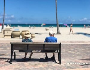 Elderly couple relaxing on a bench by the ocean, enjoying the view on a sunny beach day.