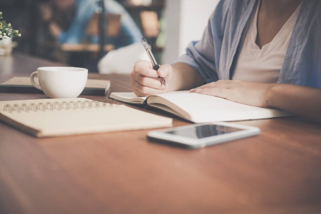 Person writing in a notebook at a desk with coffee and smartphone, symbolizing tracking progress, adjusting goals, and staying motivated with retirement planning.