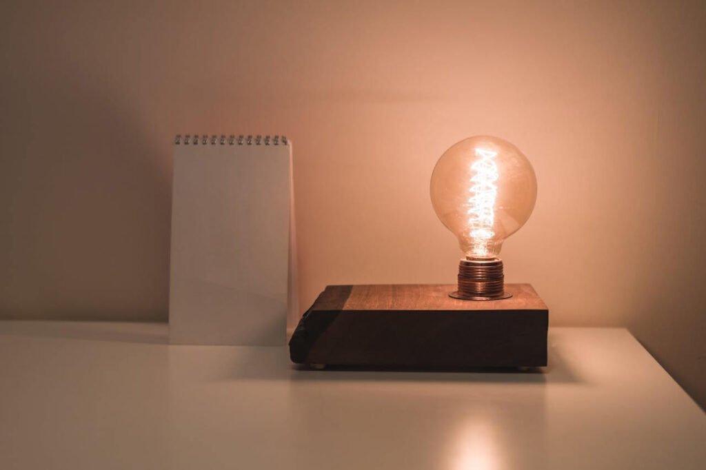 A glowing vintage-style light bulb mounted on a wooden base sits on a white desk beside a blank spiral notepad, against a softly lit beige wall.