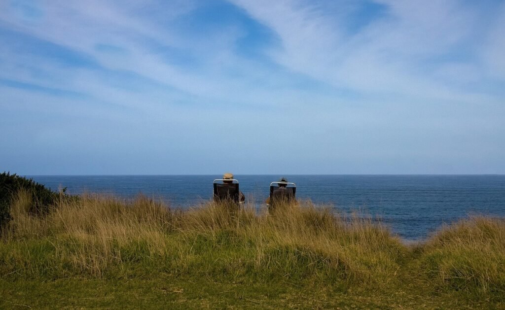 Two people sitting in outdoor chairs on a grassy hill, looking out at the ocean horizon.